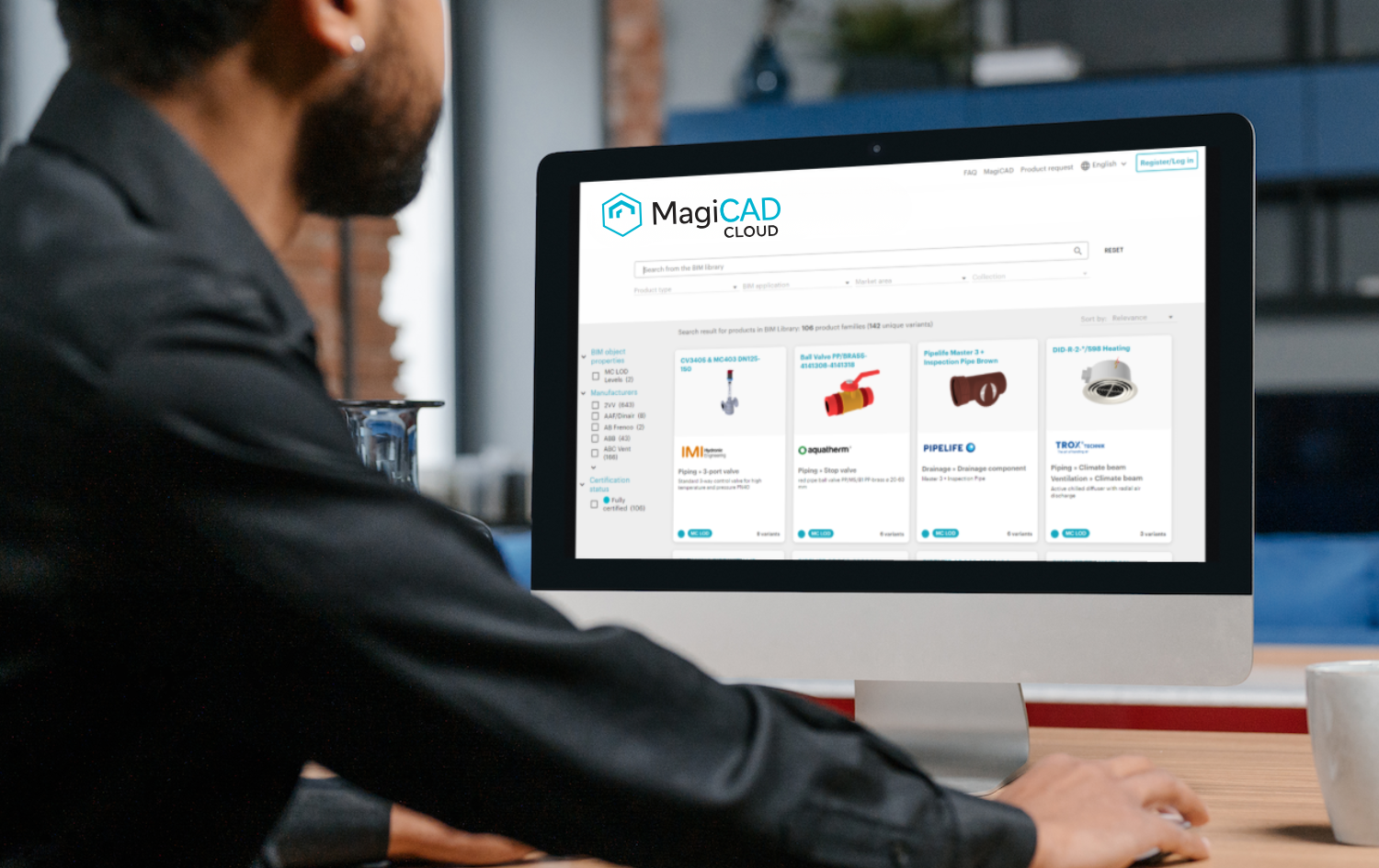 A person sitting at a desk looks at a computer screen displaying the MagiCAD Cloud website, showing BIM objects from various manufacturers with product images, filters and search options